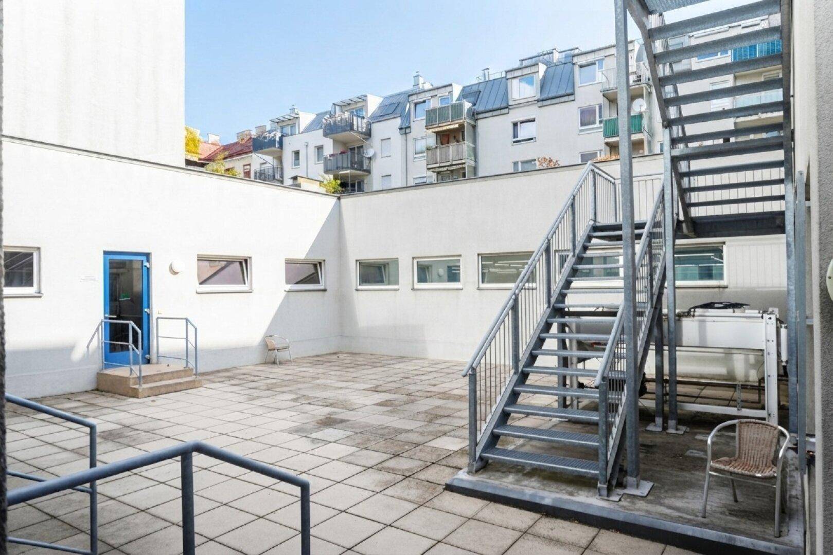 Große gepflasterte Terrasse mit Metalltreppe und Blick auf umliegende Wohngebäude in der Stadt.