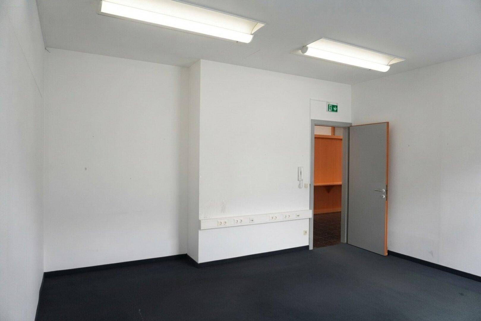 Einfacher Büroraum mit dunklem Teppichboden und weißer Wand, mit Zugang zu einem kleinen Nebenraum.