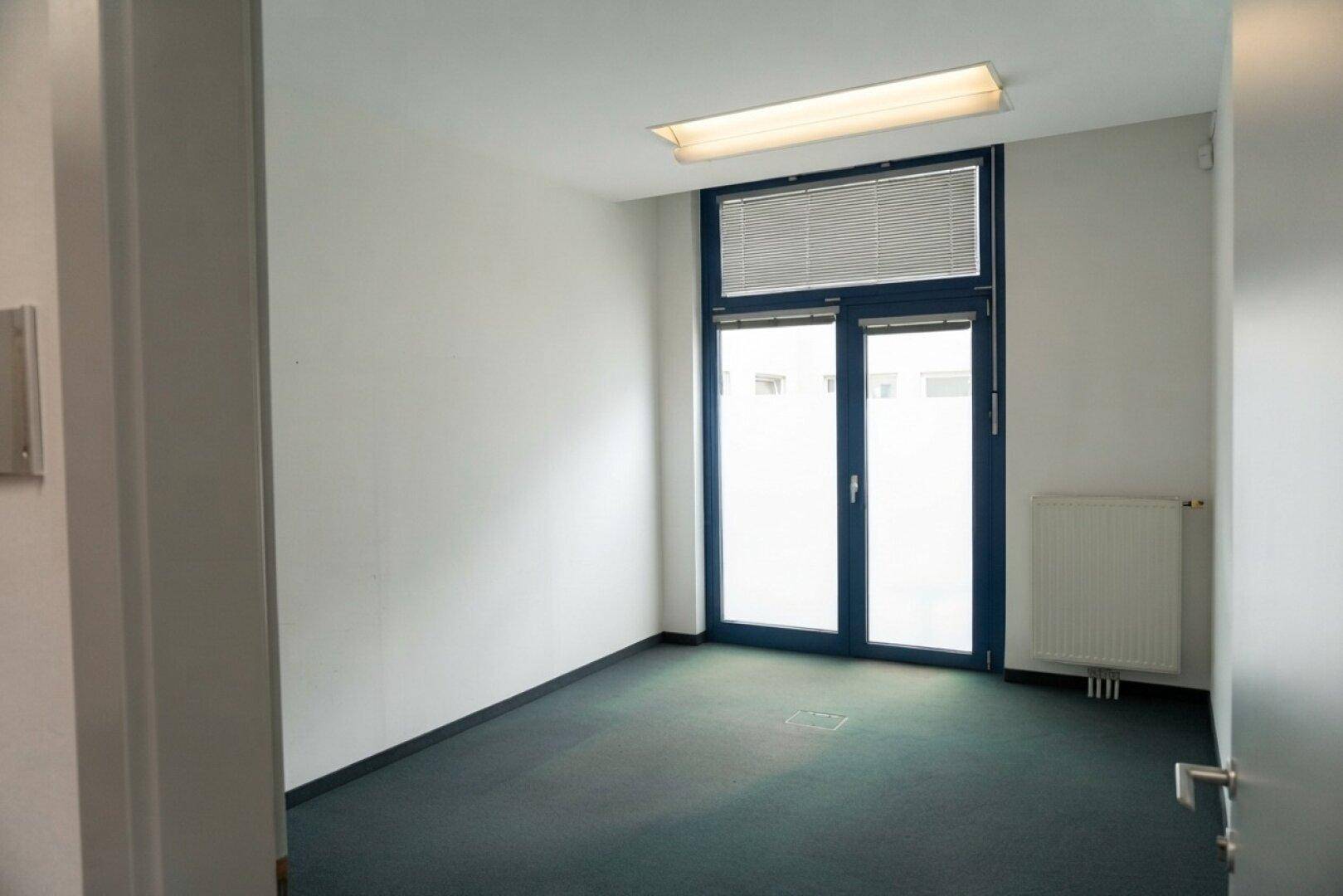 Kleiner Büroraum mit dunklem Teppichboden und Fenster mit Jalousien, bietet ausreichend Tageslicht.