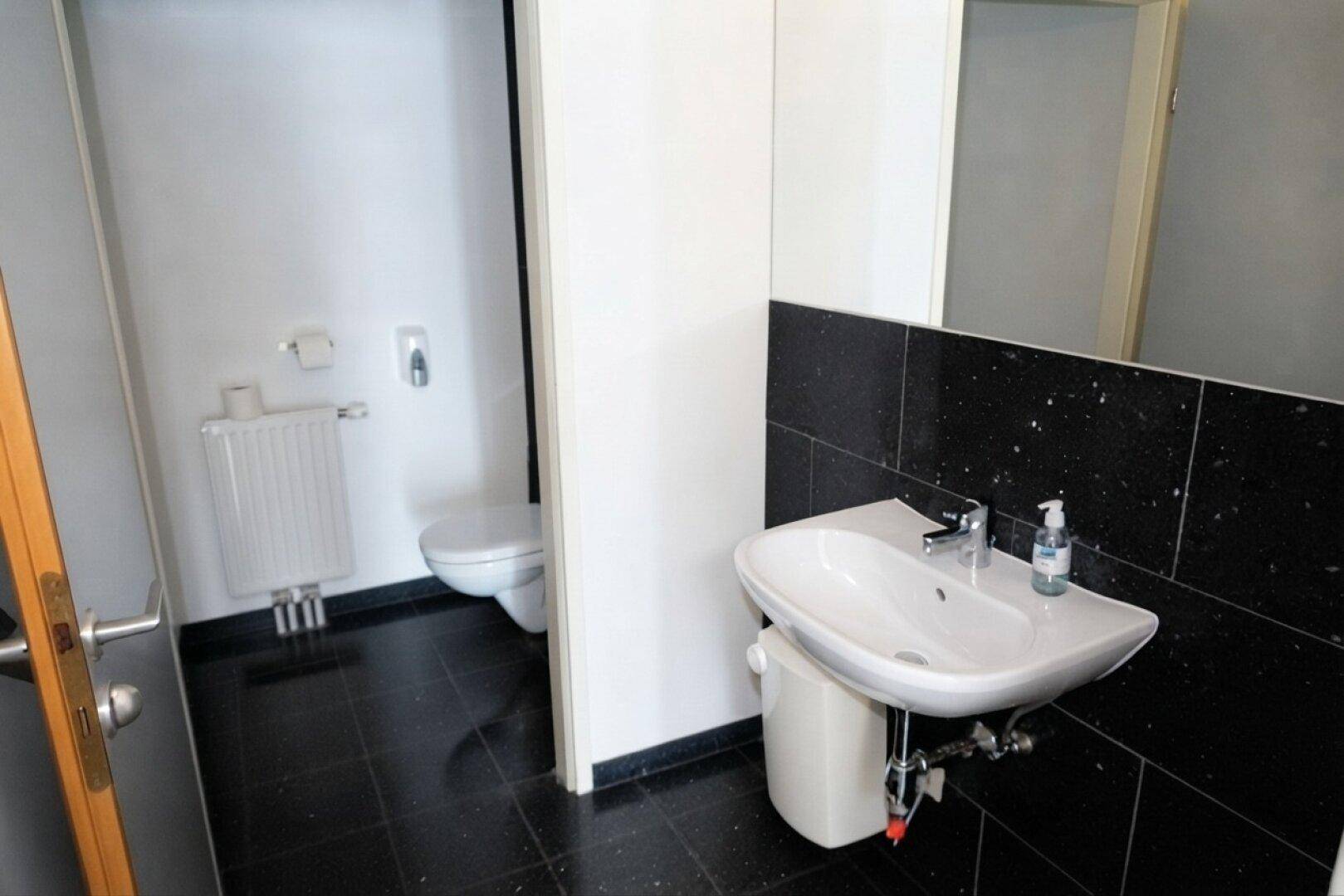 Toilettenbereich mit schwarzem Fliesenboden und Waschtisch, angrenzend an eine separate WC-Kabine.