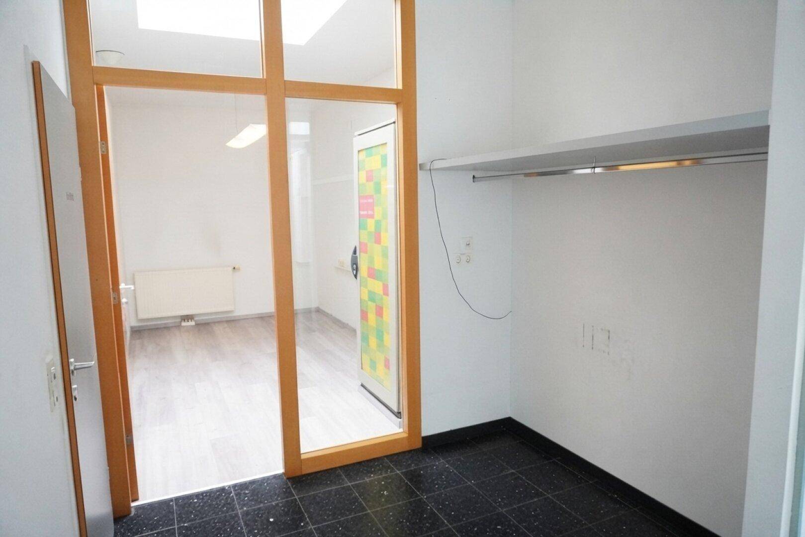 Kleiner Bereich mit dunklem Fliesenboden und einer Kleiderstange, angrenzend an Büroräume.