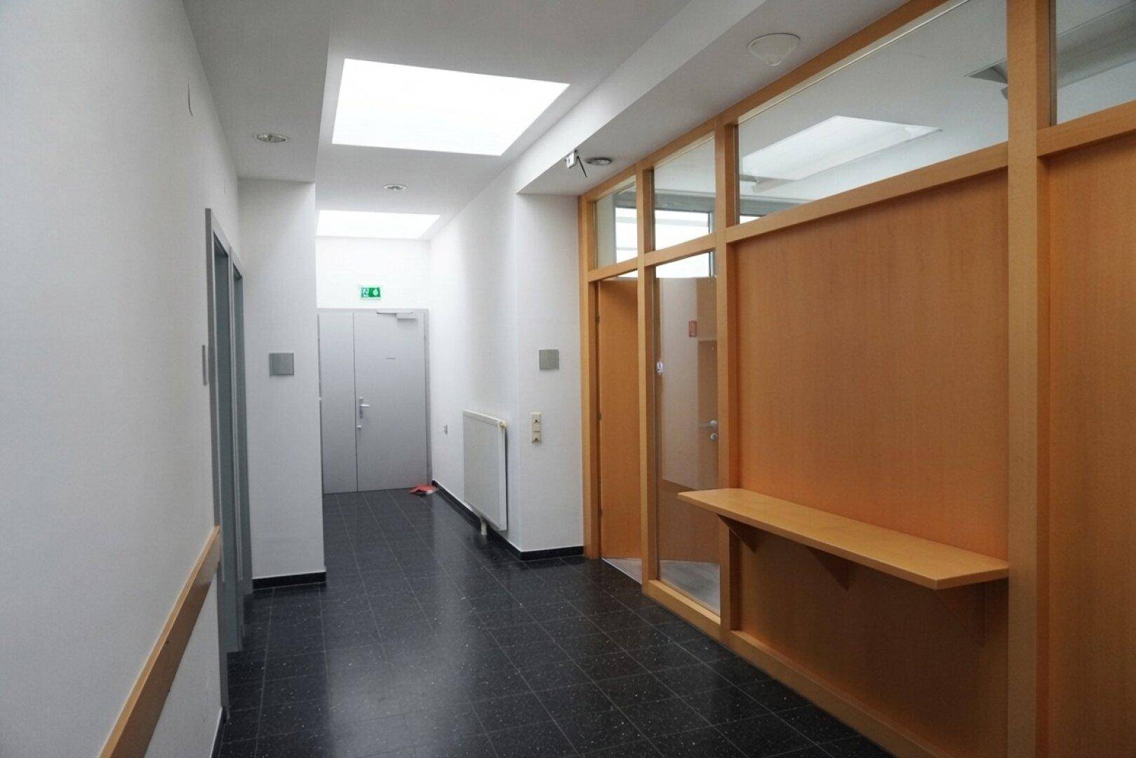 Langer, heller Flur mit Holz-Glas-Trennwänden und dunklem Fliesenboden, gut beleuchtet und sicher.