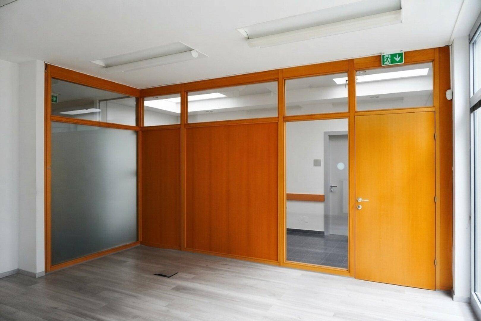 Geräumiges Büro mit hellem Laminatboden und Holz-Glas-Trennwänden, mit Zugang zum Flur.