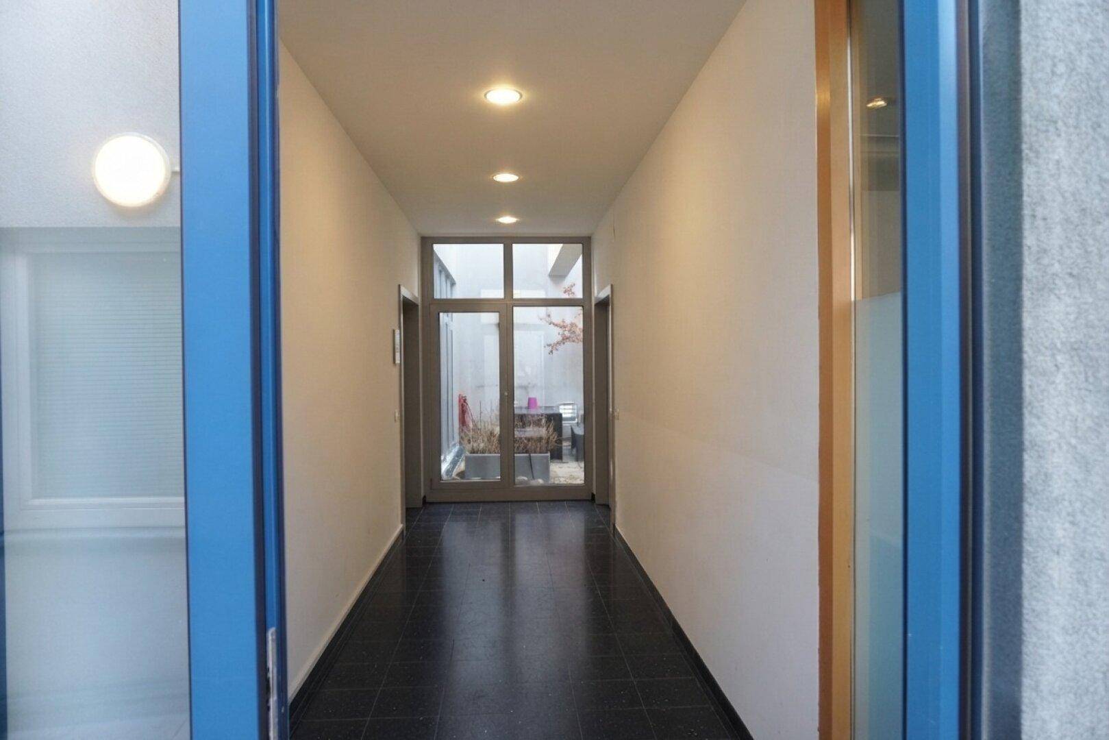 Langer, schmaler Flur mit dunklem Fliesenboden und Einbaustrahlern, führt zu einem Innenhof.