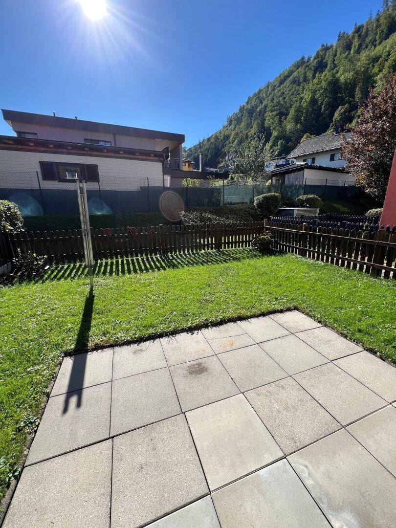 Sonniger Außenbereich mit gepflasterter Terrasse und Rasenfläche, umgeben von einem Holzzaun.