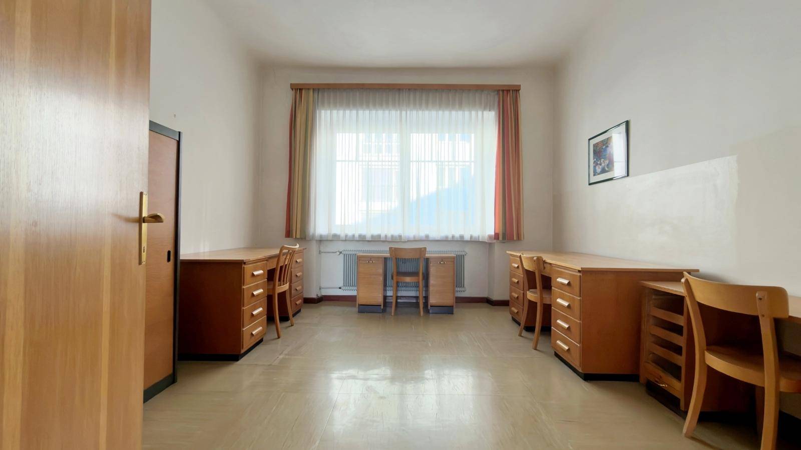 Großer Büroraum mit mehreren Schreibtischen und Stühlen, einem Fenster und hellem Bodenbelag.