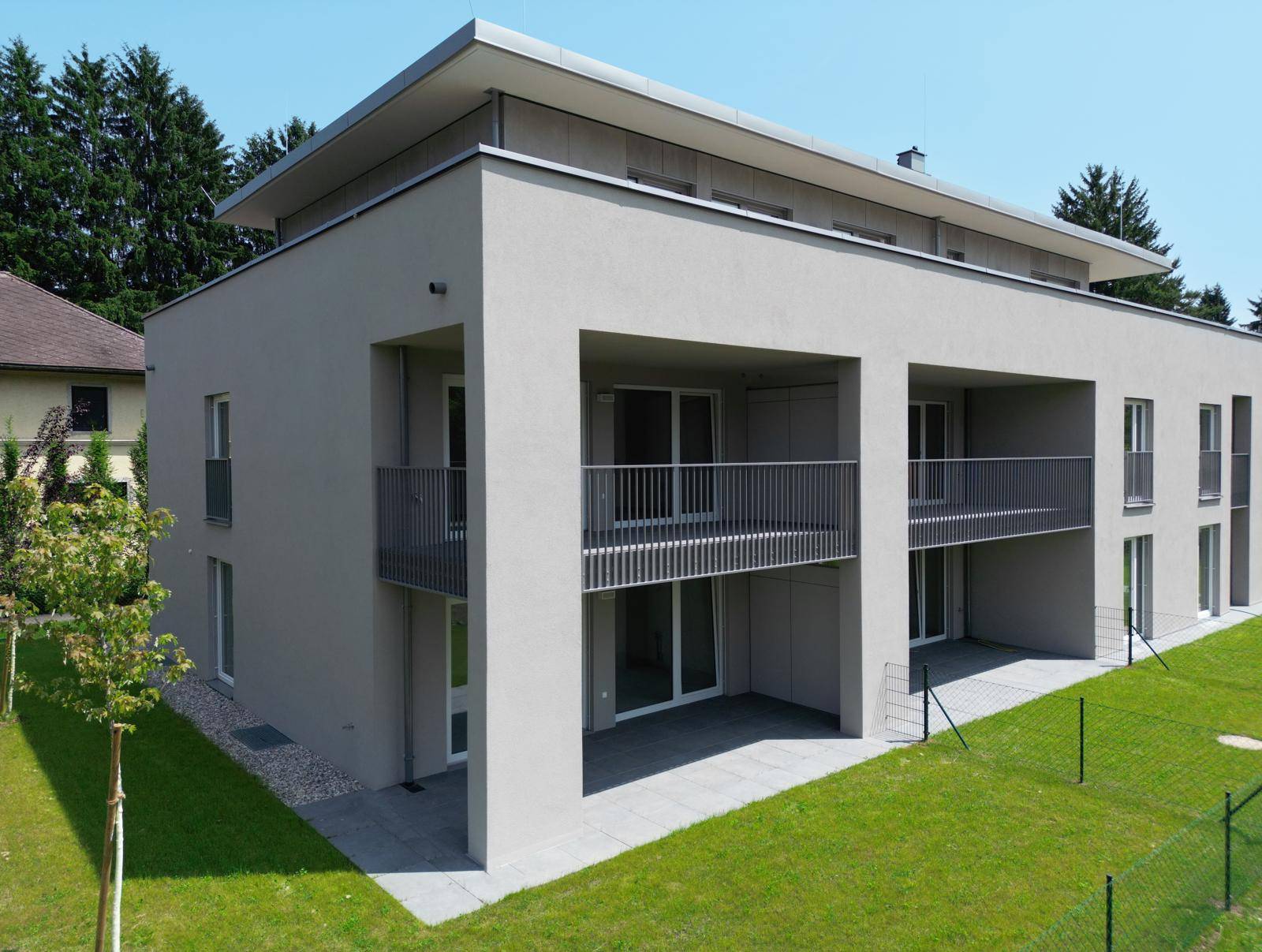 Eckansicht des modernen Wohnhauses mit hellen Fassaden, Balkonen und Terrassen in grüner Umgebung.