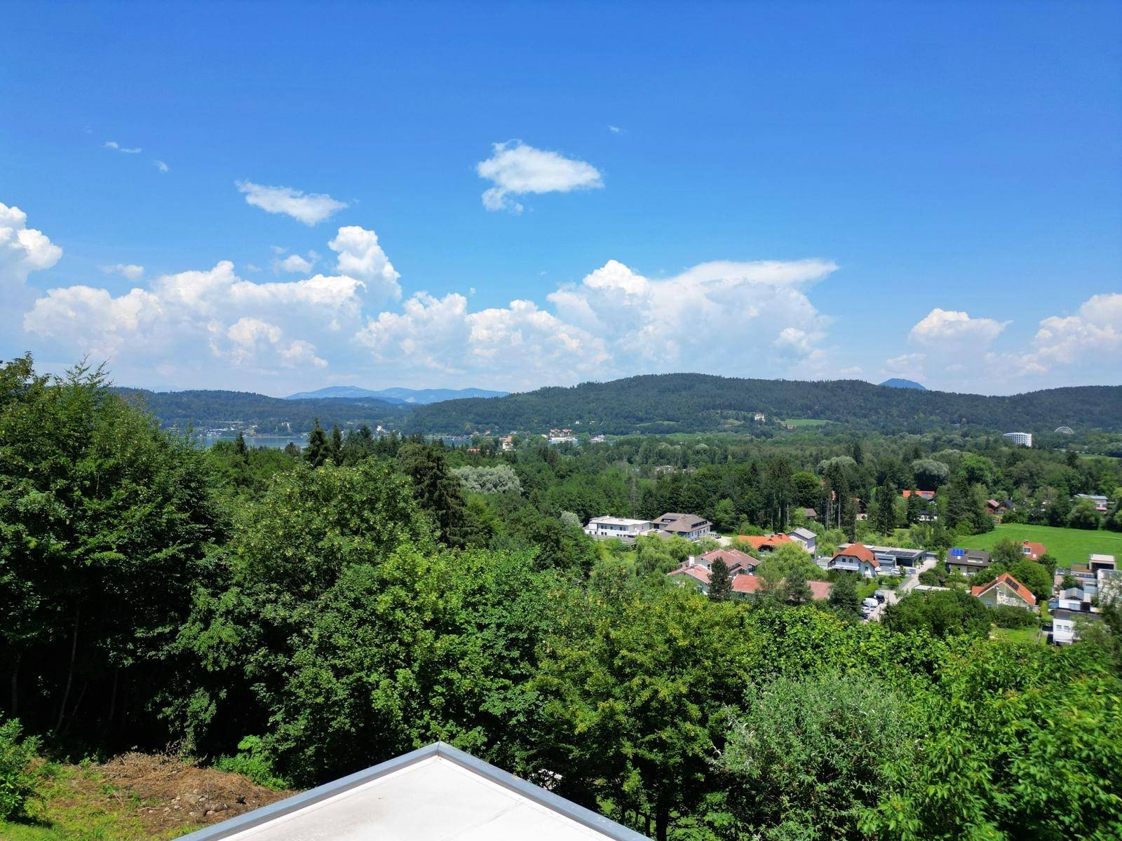 Panoramablick über eine grüne Landschaft mit See, Bergen und verstreuten Häusern unter blauem Himmel.