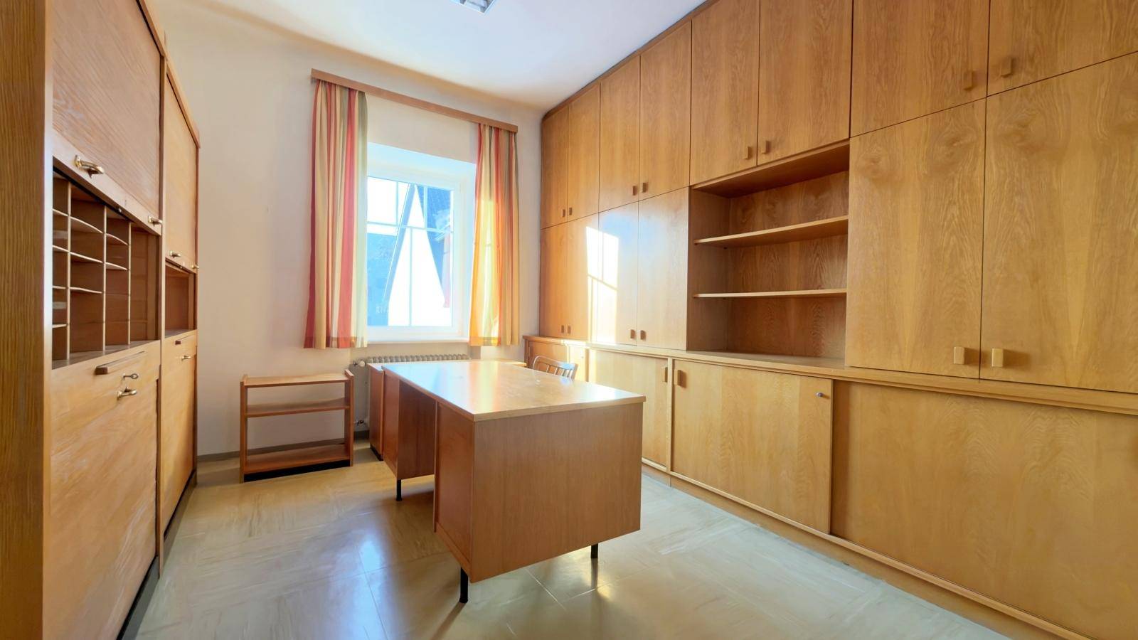 Büro mit umfangreichen Holzschränken, einem Schreibtisch und einem Fenster, das viel Licht hereinlässt.
