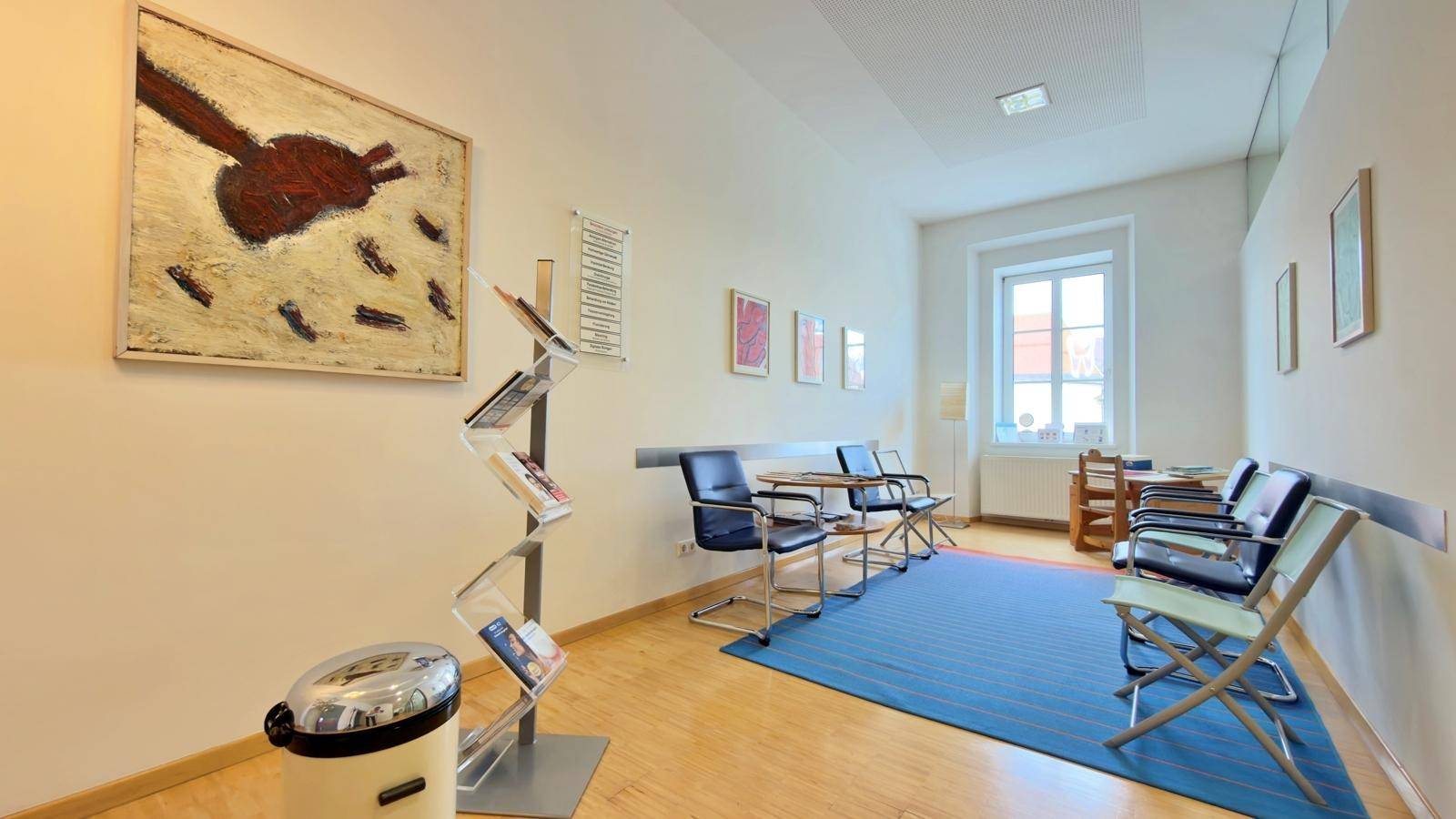 Geräumiger Wartebereich mit blauer Teppichfläche, Glaswänden und Lesematerial.