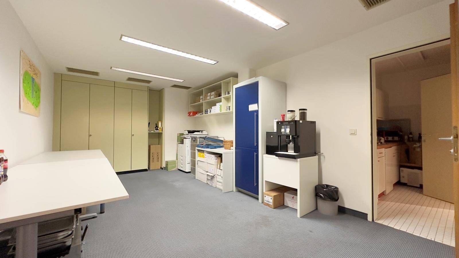 Büroraum mit Schreibtisch, Schränken, Regalen, einem Kühlschrank und einer Kaffeemaschine.