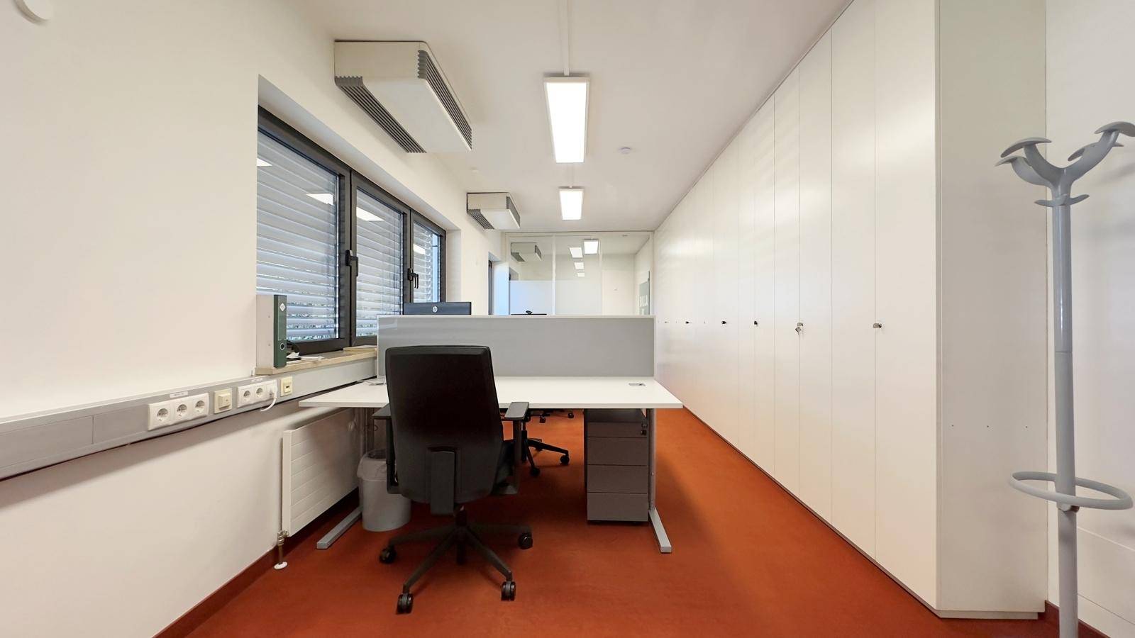 Heller Büroraum mit Schreibtischen, Bürostühlen, weißen Schränken und orangefarbenem Teppichboden.