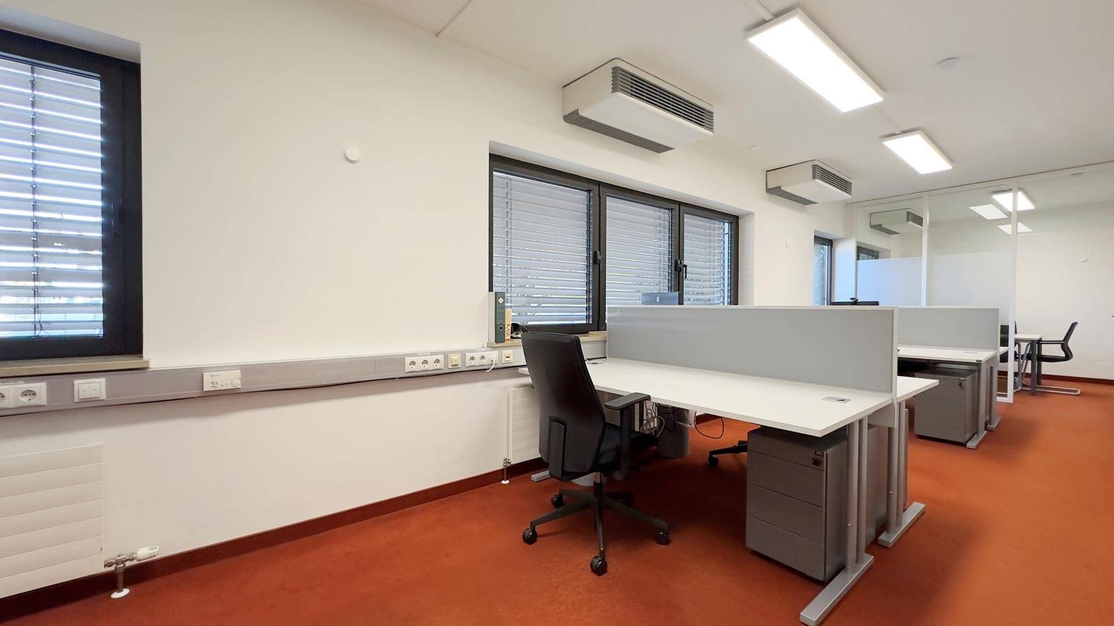 Moderner Büroraum mit mehreren Arbeitsplätzen, Fenstern mit Jalousien und orangefarbenem Teppichboden.