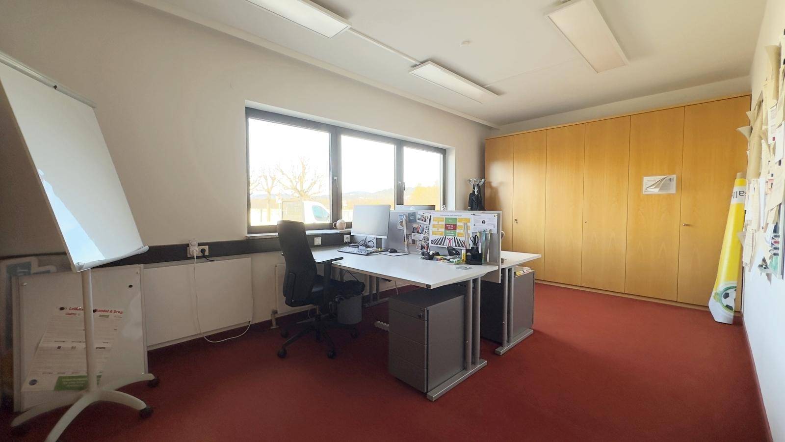 Heller Büroraum mit Schreibtischen, Bürostühlen, Holzschränken und einem Whiteboard.