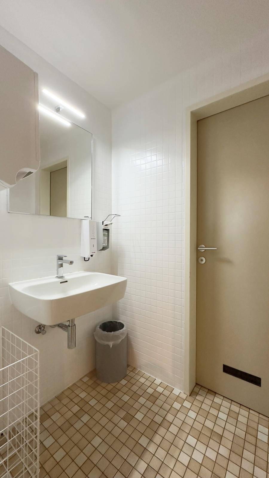 Modernes Badezimmer mit weißem Waschbecken, Spiegel und kleinen quadratischen Fliesen an Wänden und Boden.