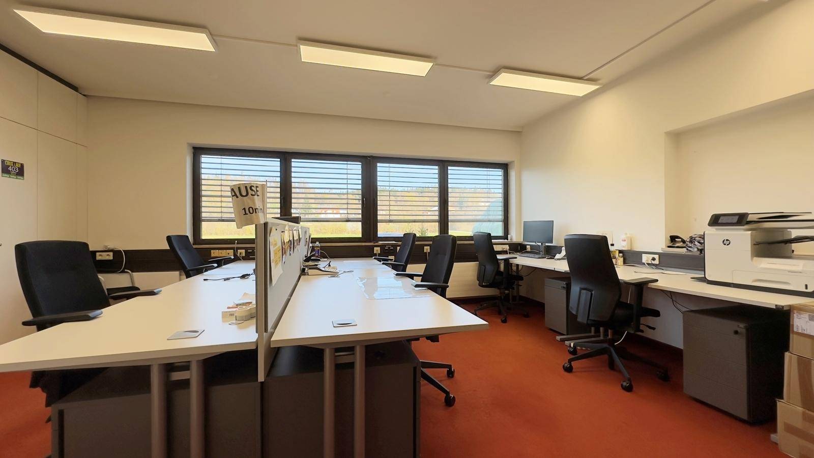 Großraumbüro mit mehreren Arbeitsplätzen, Fenstern mit Jalousien und orangefarbenem Teppichboden.