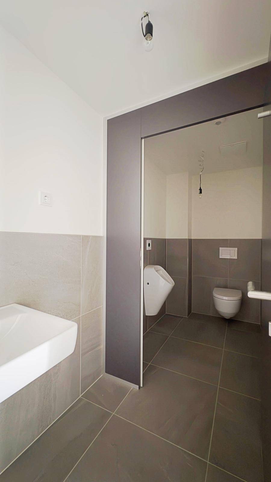 Modernes Badezimmer mit Urinal, Toilette, Waschbecken und grauen Wand- und Bodenfliesen.