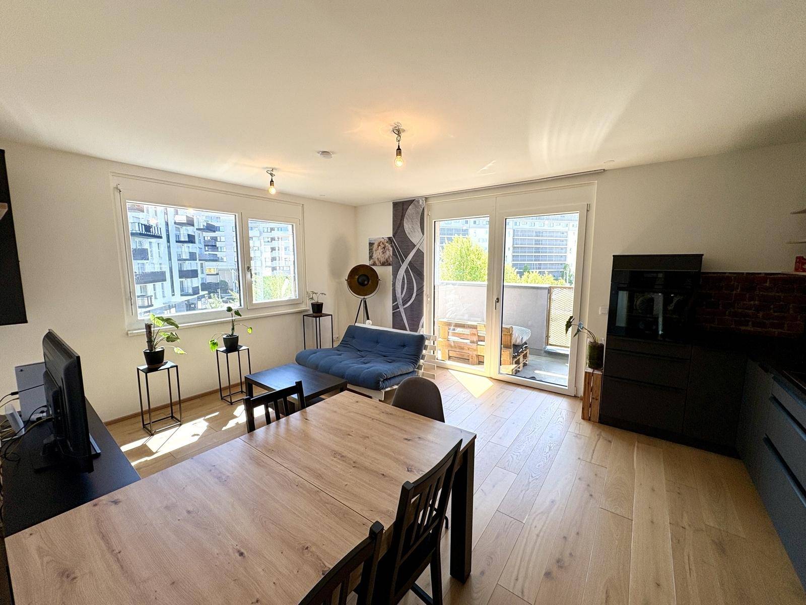 Heller Wohn- und Essbereich mit Holzboden, großem Esstisch und Zugang zum Balkon.