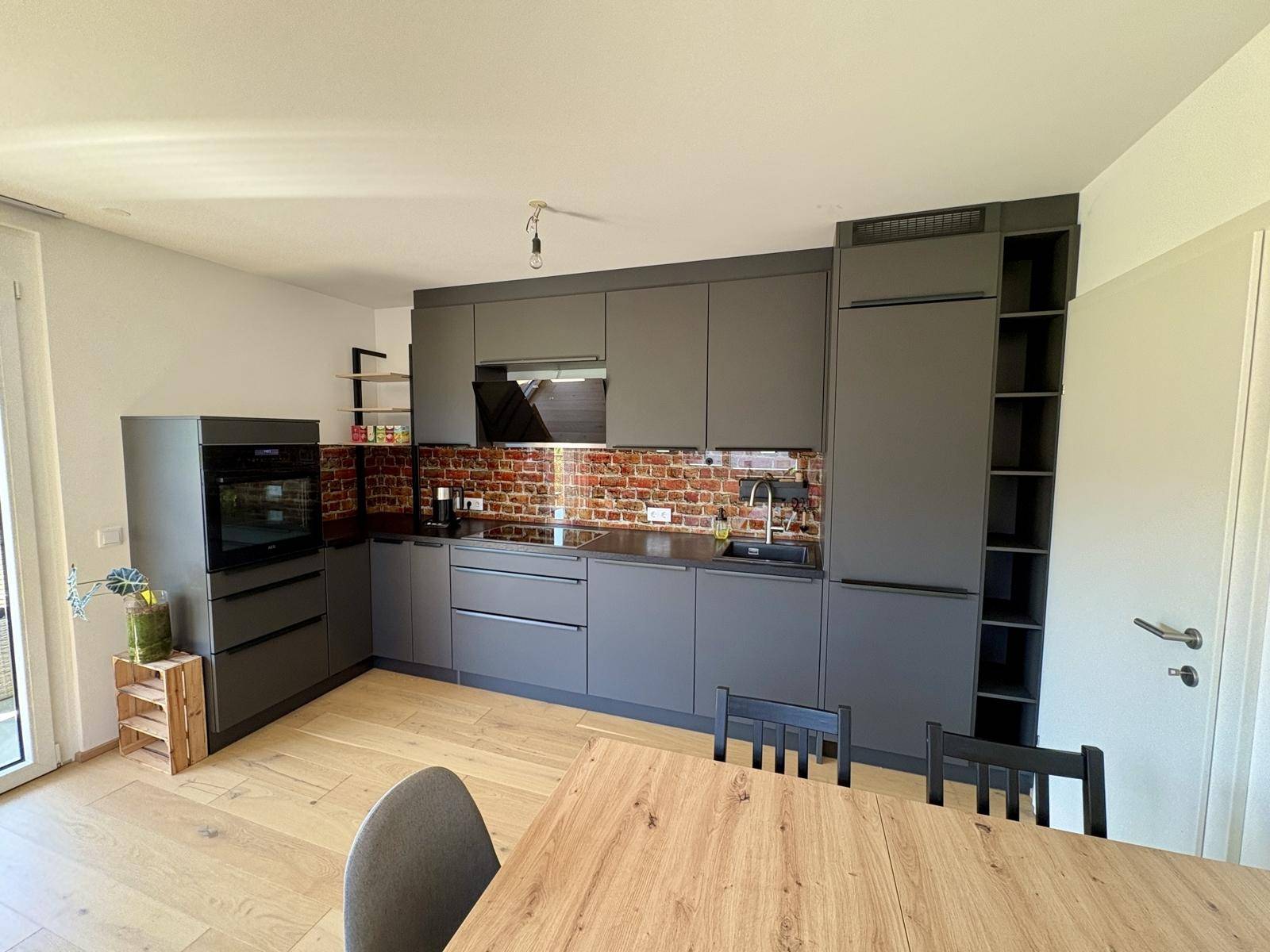 Moderne Einbauküche in Grau mit Backstein-Rückwand und Holzesstisch im offenen Bereich.