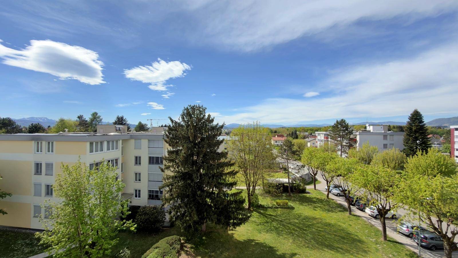 Panoramablick auf Wohngebäude, gepflegte Grünflächen und die ferne Bergkette unter blauem Himmel.