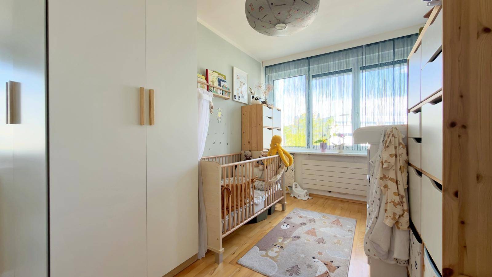 Kinderzimmer mit Babybett, hellem Schrank und großem Fenster für viel Tageslicht.
