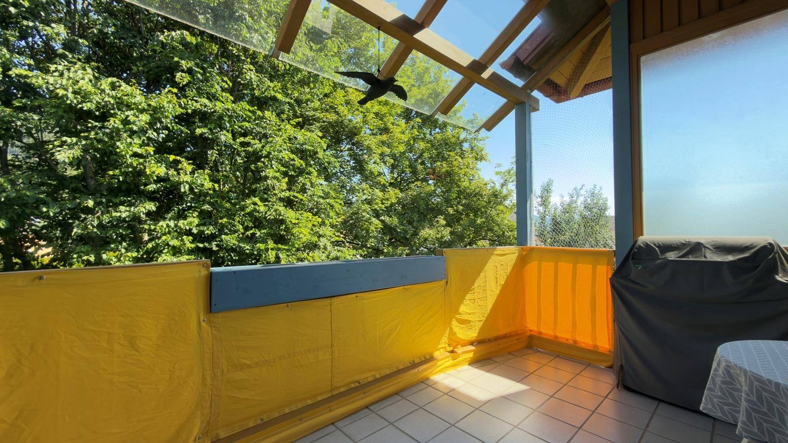 Überdachter Balkon mit Glasdach, Fliesenboden und Blick ins Grüne.