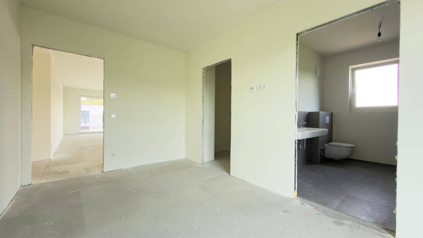 Rohbauzimmer mit Betonboden und Blick in ein teilweise fertiggestelltes Badezimmer.