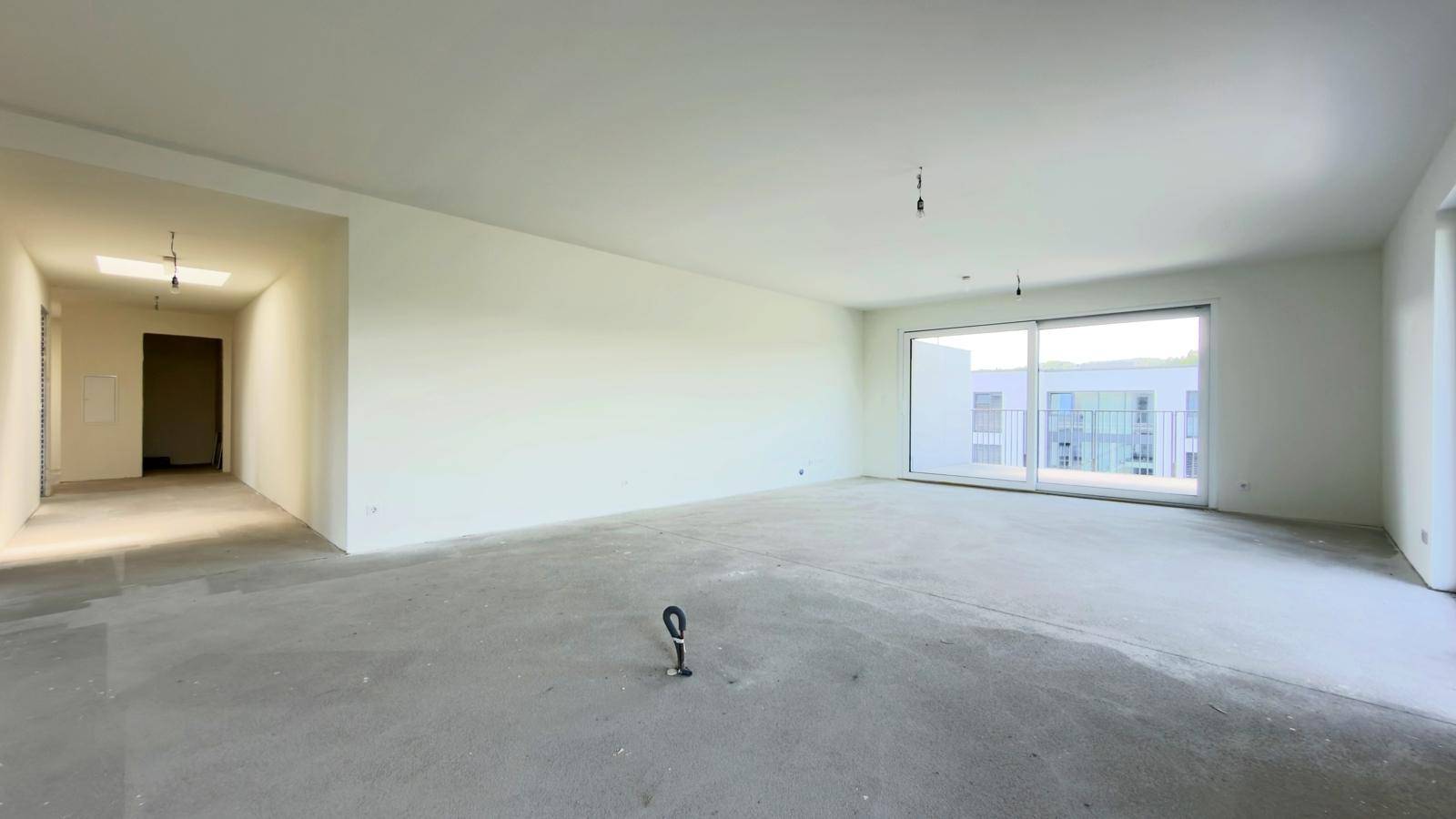 Geräumiger Wohnbereich im Rohbau mit Betonboden und viel Tageslicht durch große Fenster.