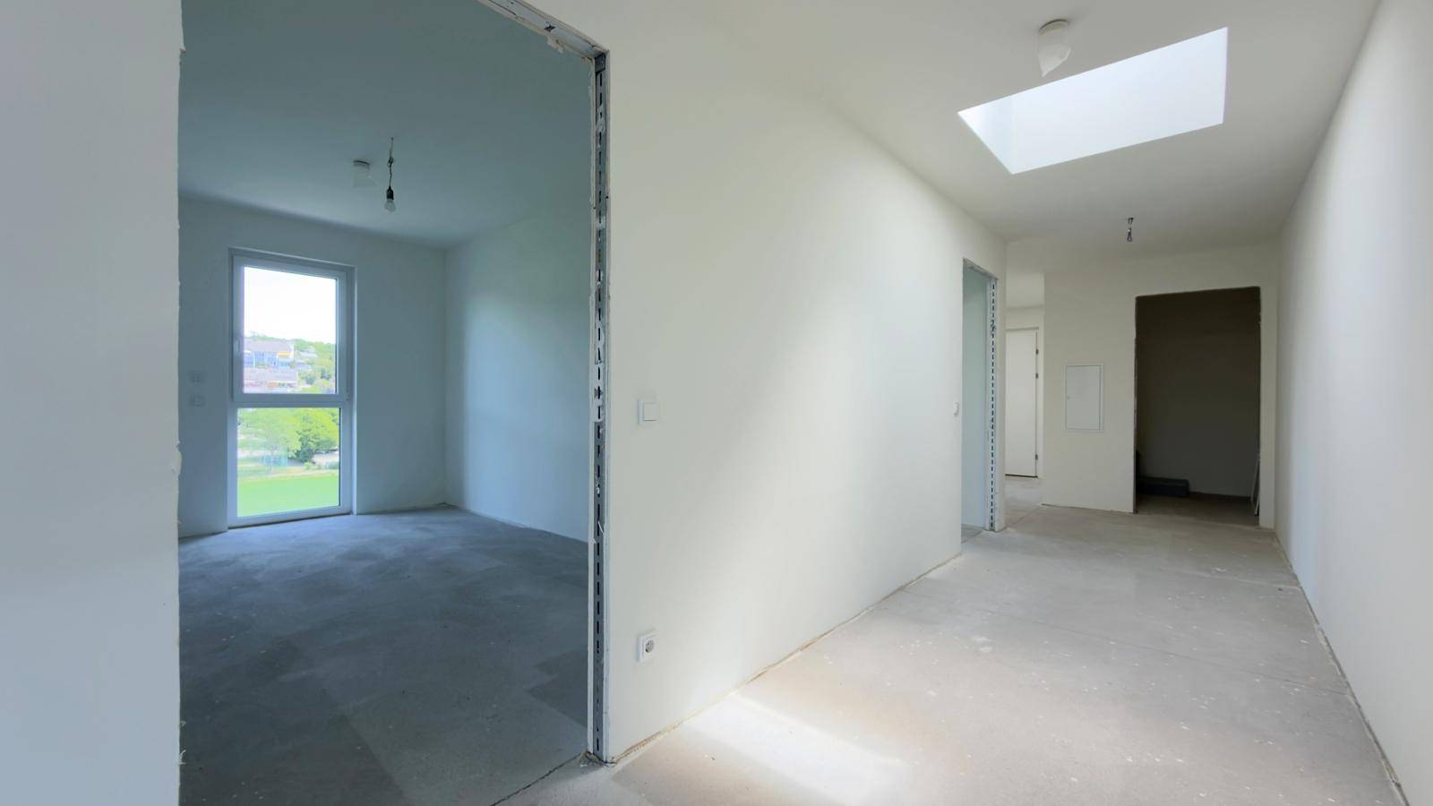 Langer Flur im Rohbau mit Oberlicht, Betonboden und Blick in ein angrenzendes Zimmer.