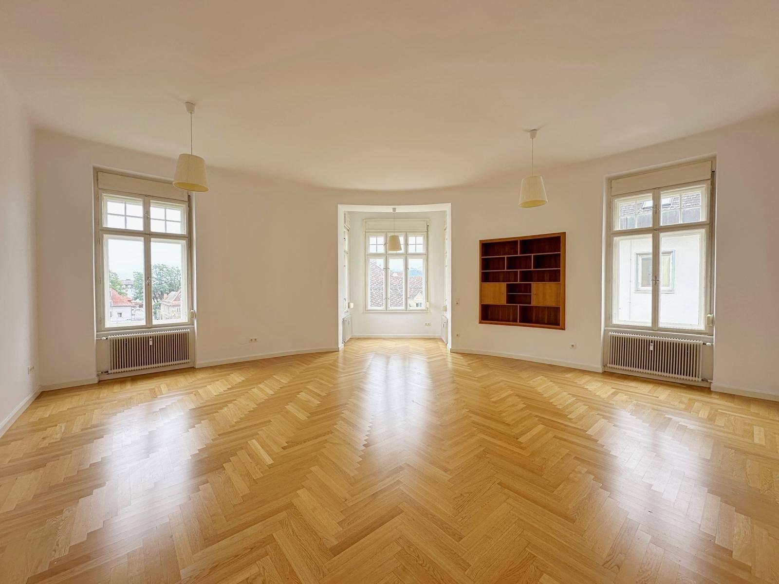 Geräumiges Schlafzimmer mit hellem Fischgrätparkett, hohen Decken und vielen Fenstern.