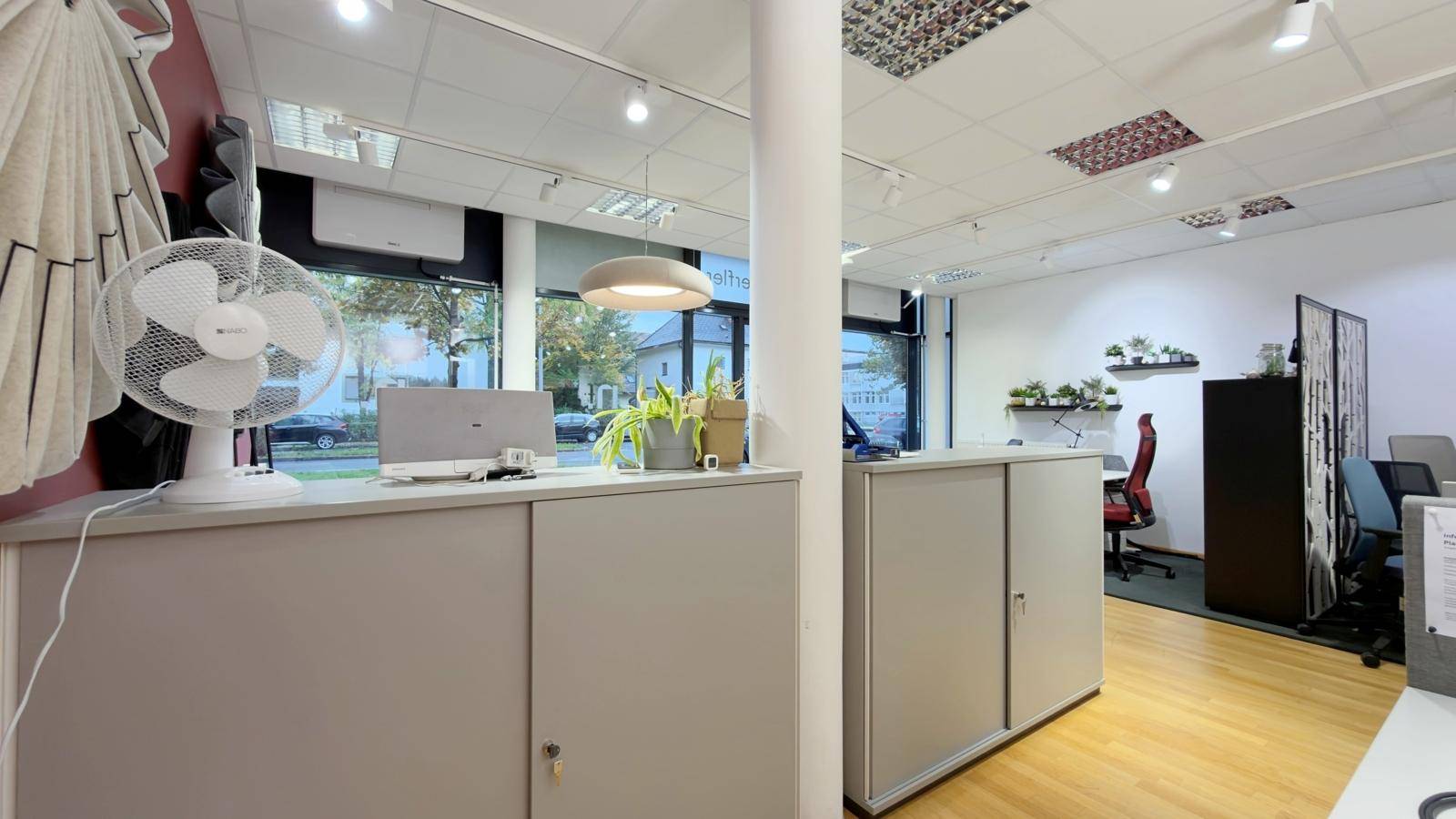 Bürobereich mit praktischen Schränken, einem Ventilator und großen Fenstern mit Blick auf die Straße.