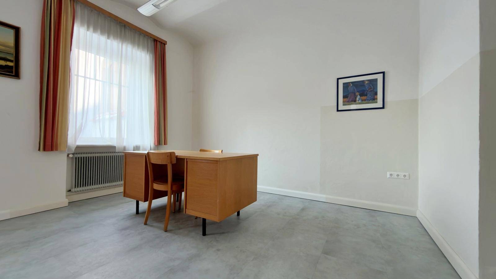 Büroraum mit einem Schreibtisch, Stühlen, großem Fenster und hellem Bodenbelag.