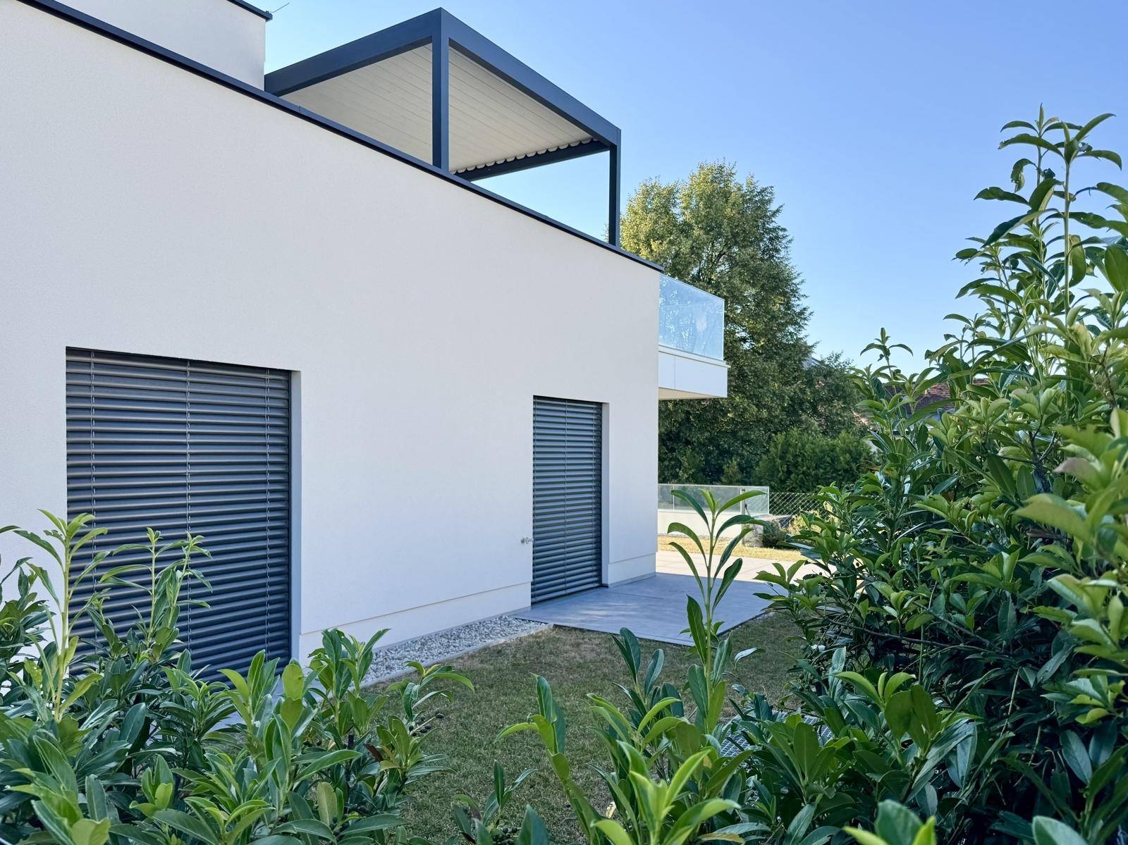 Modernes Gebäude mit weißer Fassade, dunklen Fensterläden und einer Pergola, umgeben von grünen Büschen.
