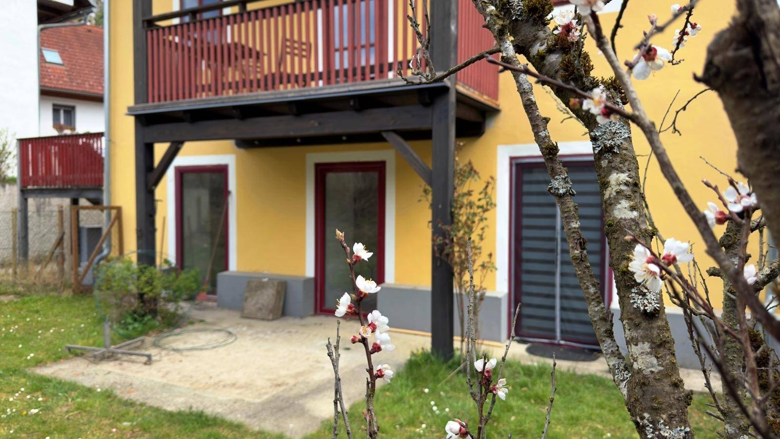 Gelbes Gebäude mit rotem Balkon und einem Gartenbereich im Frühling mit blühenden Bäumen.