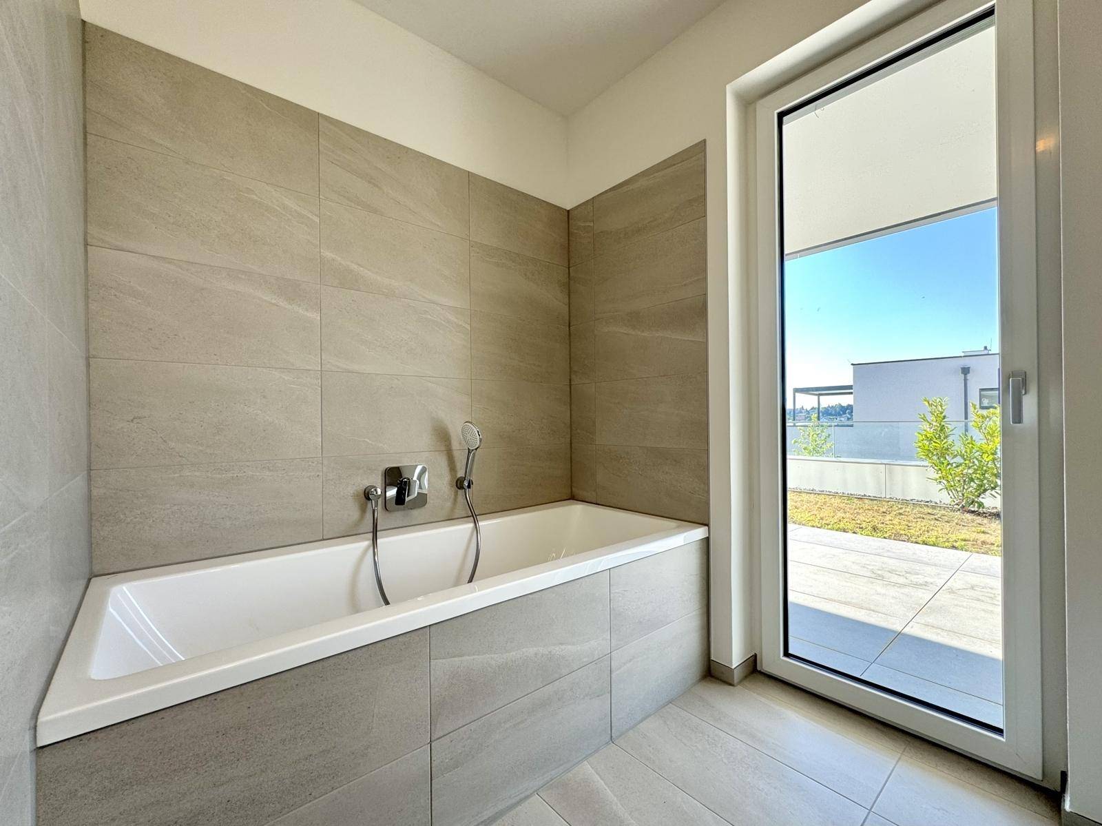 Modernes Badezimmer mit Badewanne, hellen Fliesen und Zugang zu einem Außenbereich.