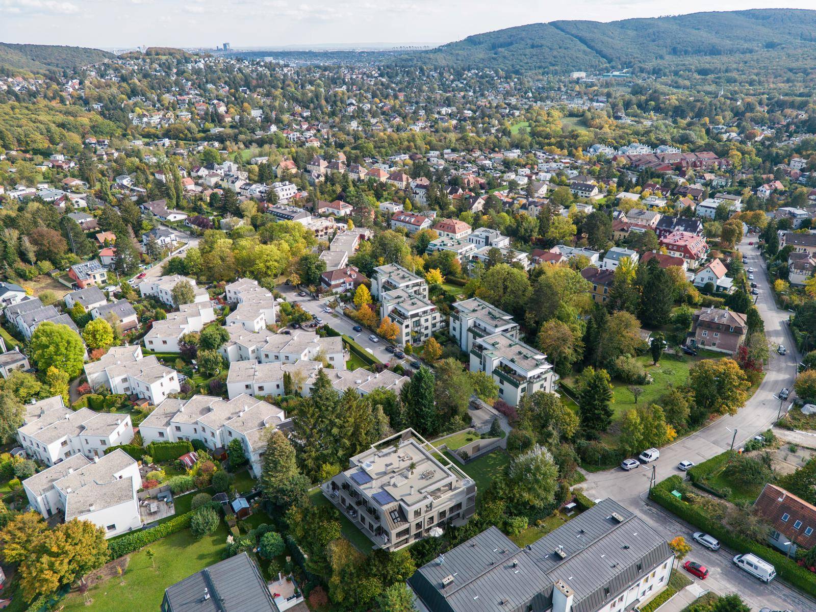 Luftaufnahme einer Wohngegend mit vielen Häusern und viel Grün, eingebettet in eine hügelige Landschaft.