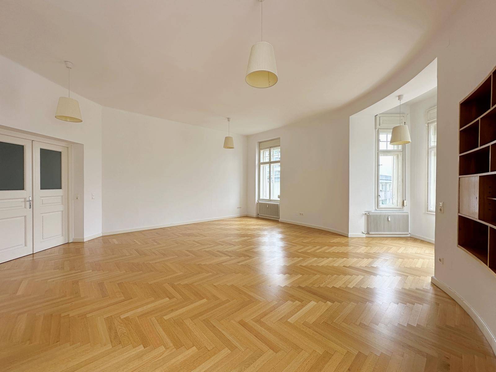 Heller, leerer Raum mit Fischgrätparkett, hohen Decken und einer markanten Rundung in der Wand.