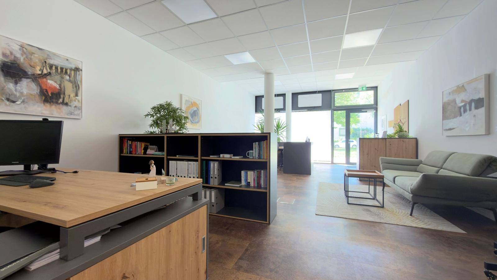 Geräumiges und helles Büro mit modernen Möbeln, Regalen und einem Wartebereich mit Sofa.