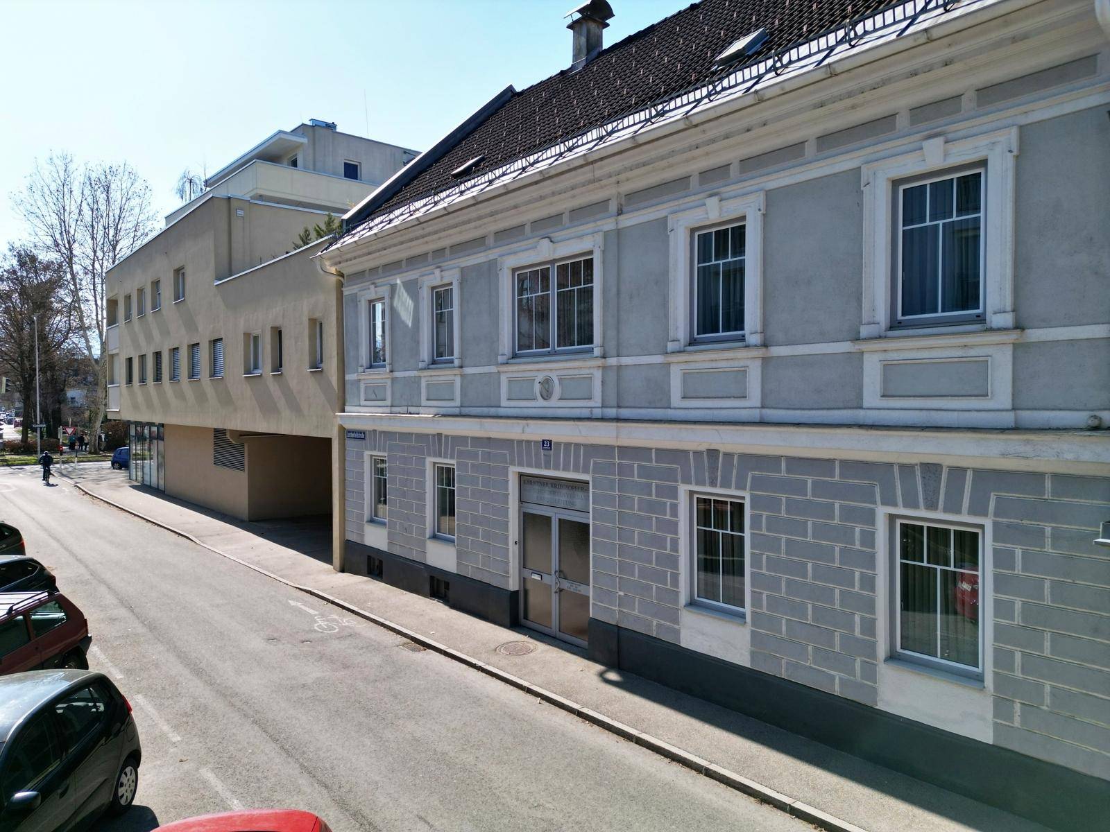 Die Fassade eines älteren Gebäudes mit angrenzendem modernen Bau und parkenden Autos an der Straße.