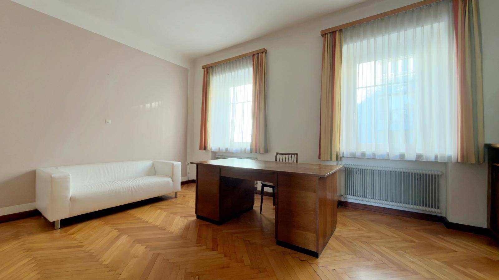 Heller Raum mit Fischgrätparkett, einem weißen Sofa und einem großen Schreibtisch vor den Fenstern.