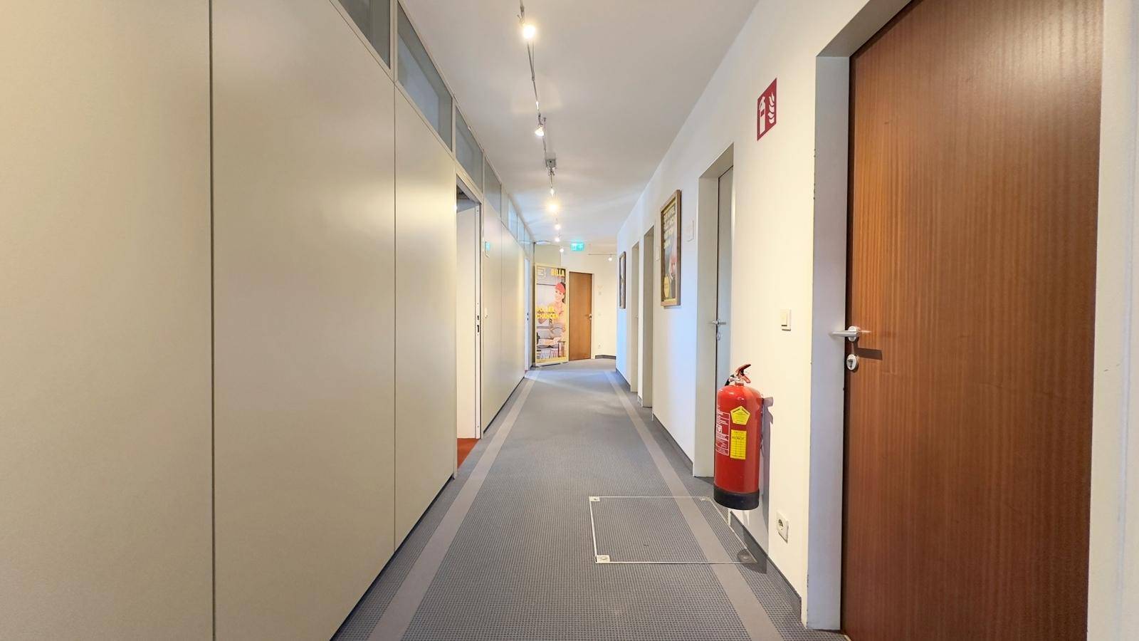 Langer, heller Flur mit Einbauschränken entlang der Wand und einem Teppichboden in Grau.