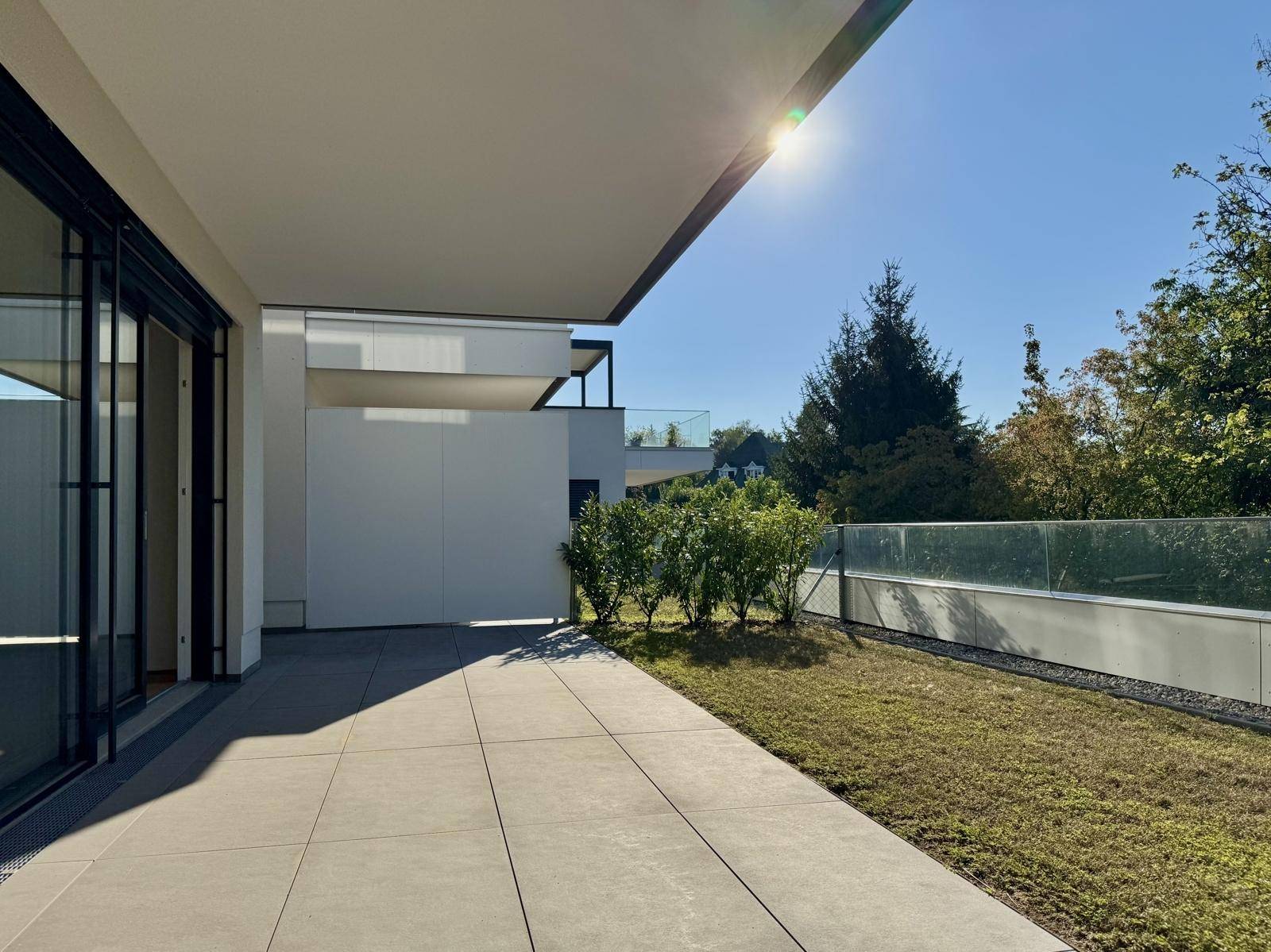 Sonniger Terrassenbereich mit Fliesenboden, kleiner Rasenfläche und moderner Glasbrüstung unter einer Pergola.