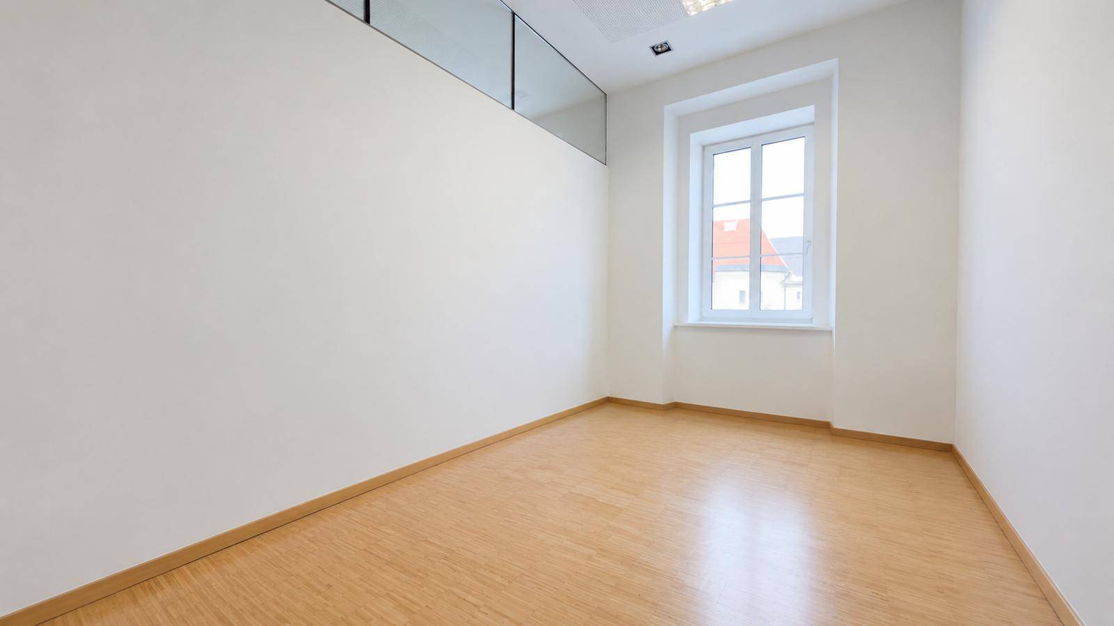 Leerer Raum mit Holzfußboden, weiß gestrichenen Wänden und einem Fenster, das Tageslicht hereinlässt.