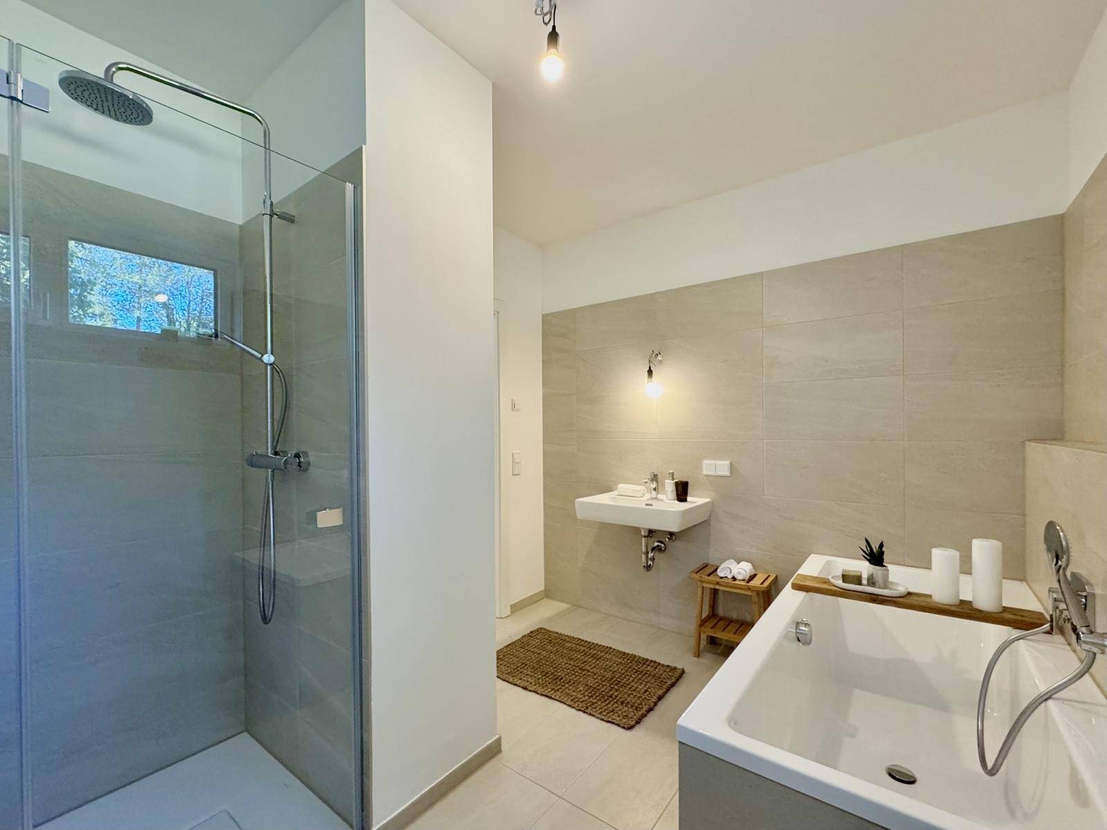 Modernes Badezimmer mit hellgrauen Fliesen, begehbarer Dusche, Badewanne und Fenster für Tageslicht.