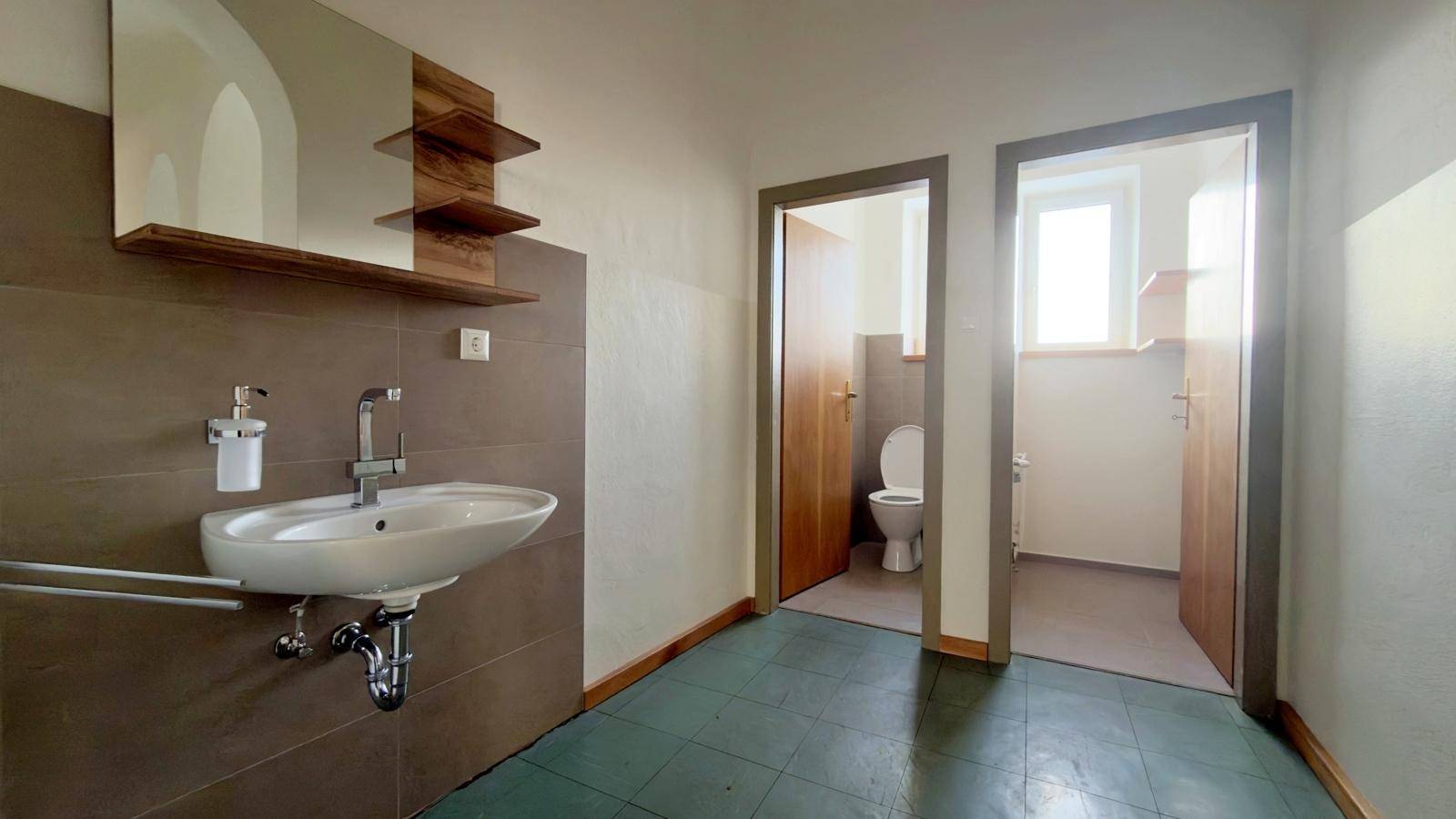 Badezimmer mit Waschbecken, Spiegel und zwei separaten Toilettenräumen, ausgestattet mit Fliesenboden.