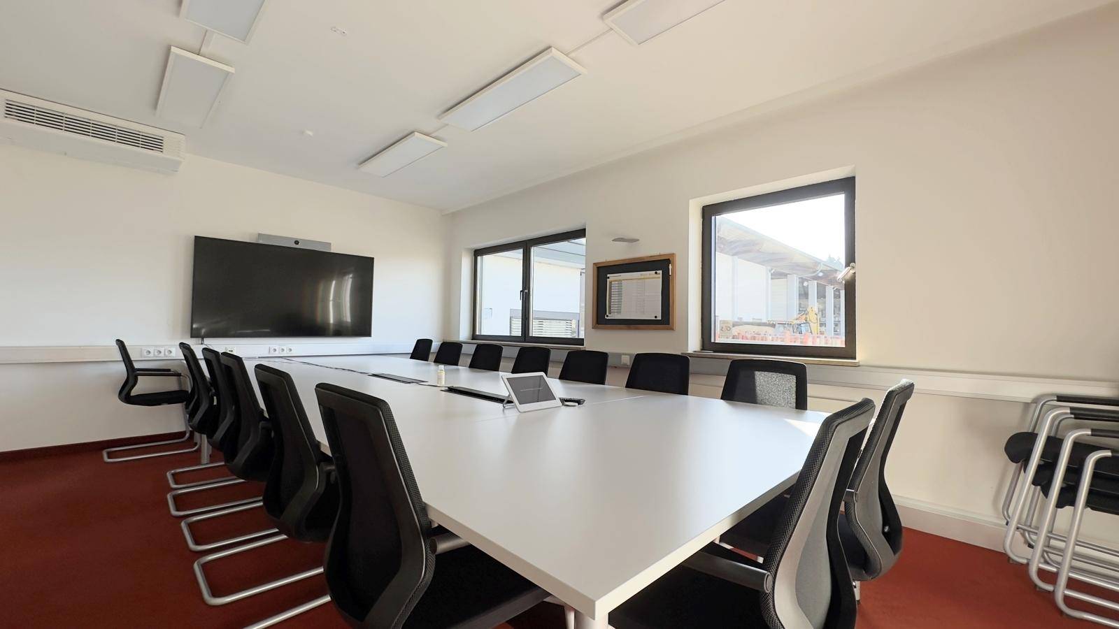 Geräumiger Konferenzraum mit langem weißen Tisch, vielen Stühlen und großem Bildschirm für Präsentationen.