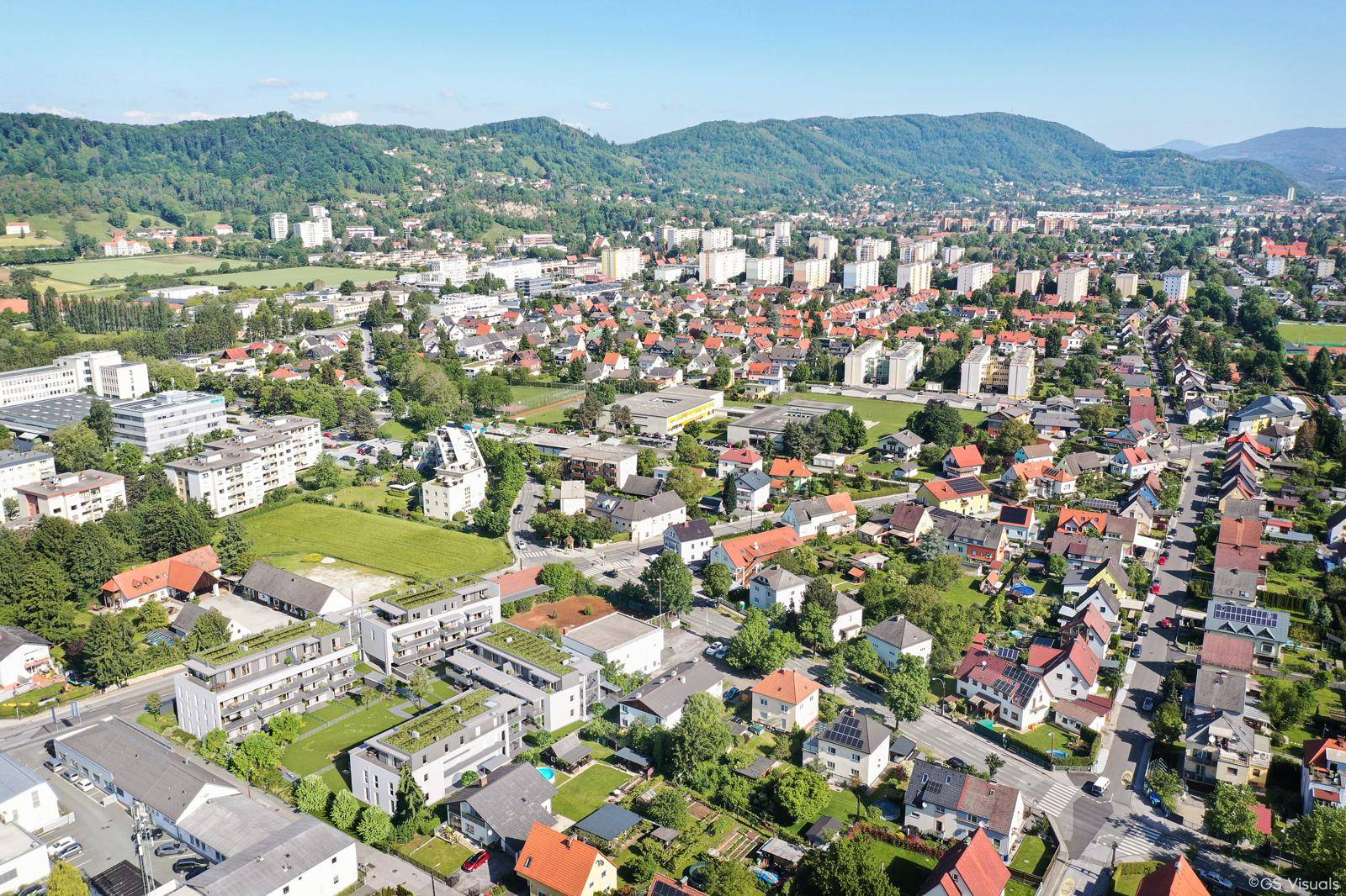 Weitläufige Luftaufnahme einer Stadt mit Wohngebieten und bewaldeten Hügeln im Hintergrund.