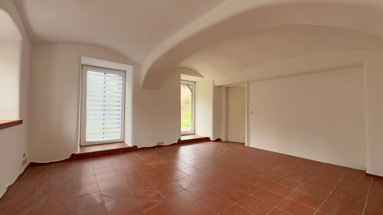 Heller Raum mit Terrakottafliesen, hohen Gewölbedecken und zwei Fenstern mit Jalousien.