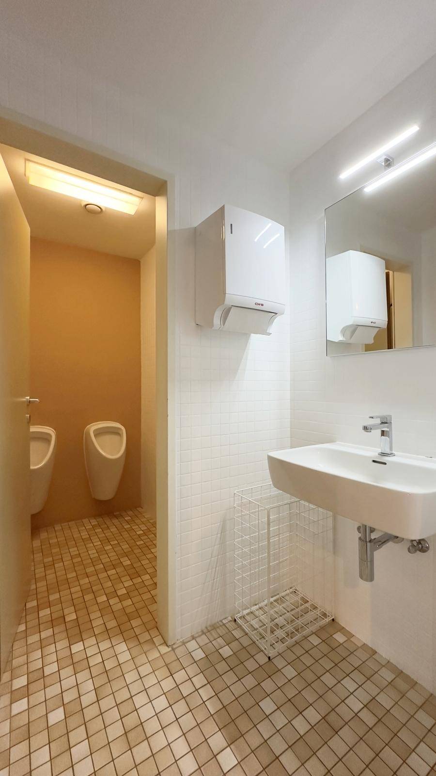 Herren-WC mit Waschbecken, zwei Urinalen und hellen Fliesen an Wänden und Boden.