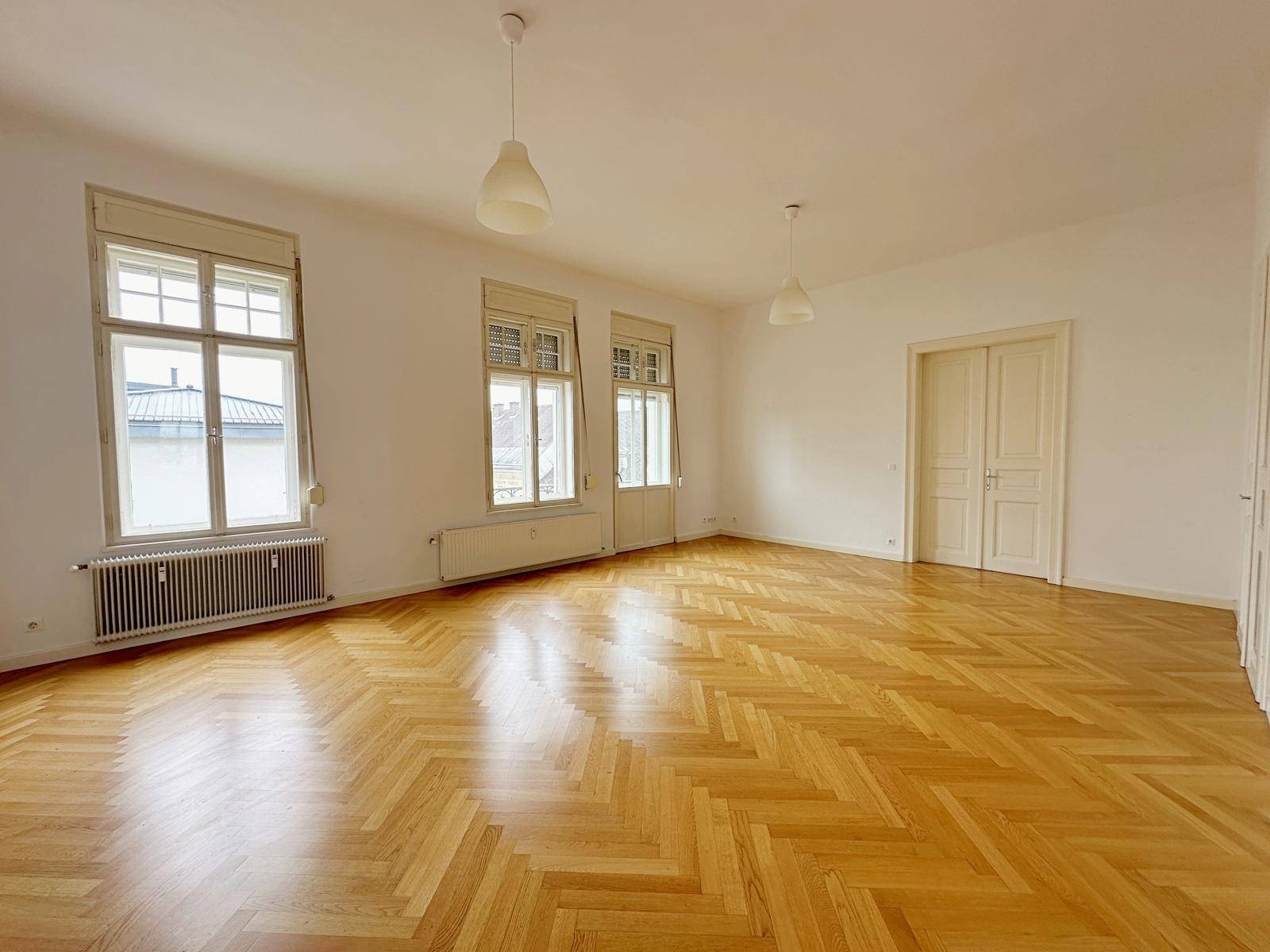Leeres Wohnzimmer mit glänzendem Fischgrätparkett, hohen Decken und großen Fenstern.